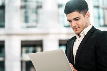 a dark-haired businessman of European appearance stands in a business center in a stylish black suit. holds in his hands works on a laptop computer. copy space. banner for the site.