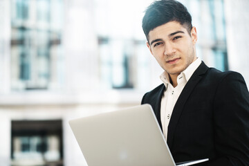 a dark-haired man of European appearance stands in a business center in a stylish black suit. holds in his hands works on a laptop computer. copy space. banner for the site.