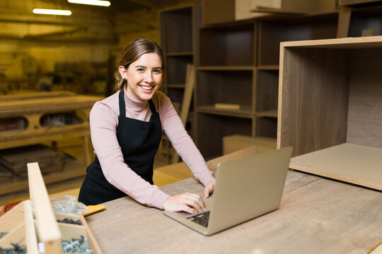 Smiling female carpenter working on a laptop in her wood workshop