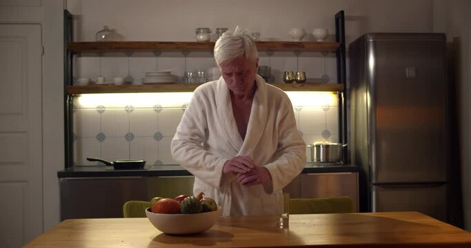 Senior Man In Bathrobe Taking Pills And Drinking Water In Kitchen In Evening