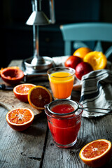 Glass of fresh blood orange juice, orange halves and juicer machine on wooden table with chair on background .