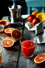 Glass of fresh blood orange juice, orange halves and juicer machine on wooden table with chair on background .