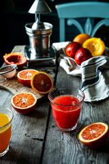 Glass of fresh blood orange juice, orange halves and juicer machine on wooden table with chair on background .