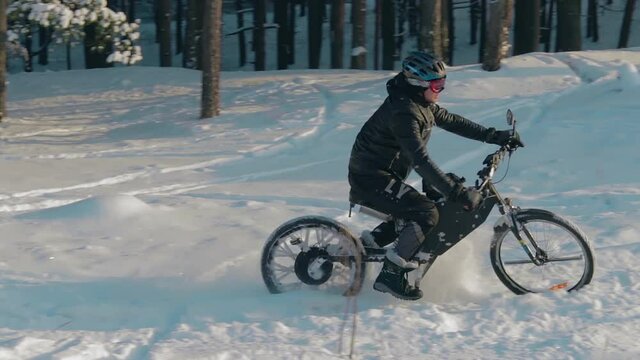 Cyclist in Black Clothes, Helmet and Snow Glasses Riding on Electric Bike in the Snow in Winter Forest. E-Bike Riding and Drifting at Cold Sunny Day - Slow Motion