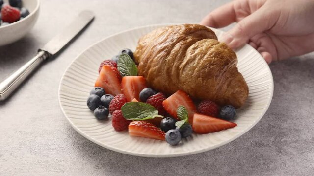 The girl puts and puts a plate with a croissant and berries on the table, a delicious breakfast 