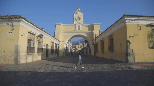 Young Hispanic woman walking in emblematic street of Antigua Guatemala below Arch of Santa Catalina - young man walking through colonial city early in the morning 