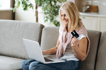 Happy middle aged smiling lady, shopping online. Joyful caucasian senior blonde woman holds the credit card, shopping online, online payment using laptop, sitting at living room