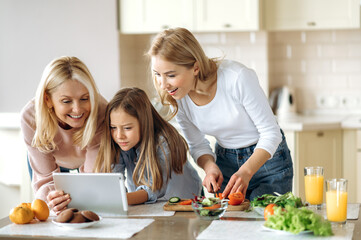 Three happy female generation at the kitchen at home. Caucasian happy grandma daughter and granddaughter cook together a fresh salad while look at the recipe on the internet, and smiling