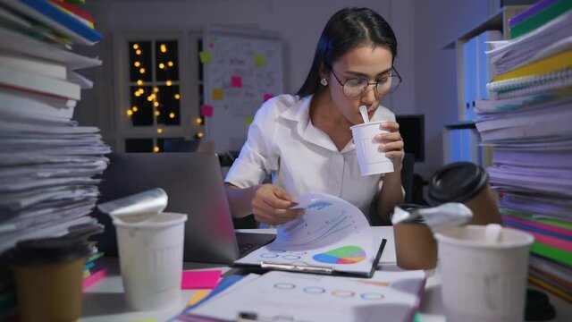 Young asian business woman is sitting at desk cover with stack of paperwork and eating instant noodles for a late meal
