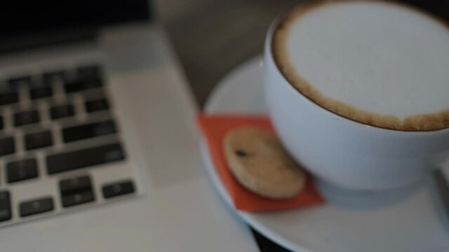 A cup of cappuccino coffee or latte coffee in a white cup with a laptop on the table. Royalty high-quality free stock footage of drink capuccino or latte coffee with a laptop for working in an office