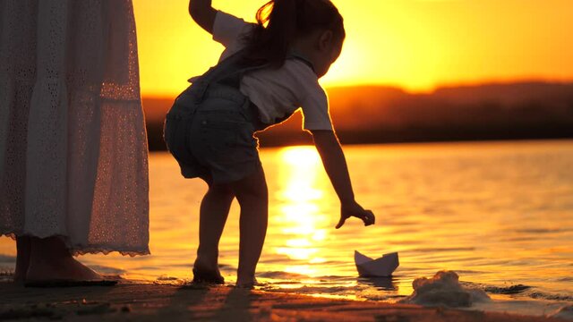 Children put a paper boat into water. Girls play with paper boats. Child's hands launches boat in park into river. Paper fleet. Happy family fantasy, childhood dream, adventure and travel concept.