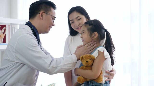 Male pediatrist touching or palpating female child patient to examine tonsils and lymph nodes, comforting and cheer up the girl. Asian mother and child see doctor concept