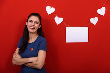 Beautiful brunette woman wearing casual blue t-shirt over isolated red background smiling near rectangular banner and white hearts