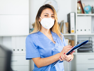 Portrait of positive latina female doctor in medical mask and blue overall writing notes on...
