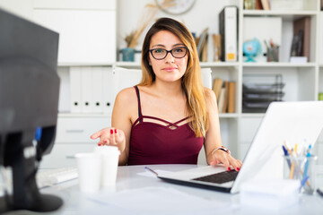 Positive latina woman in casual wear working alone with laptop and papers in office