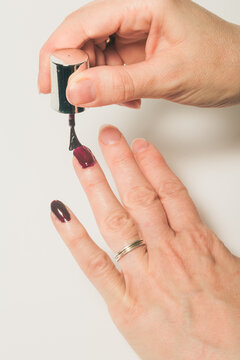 Woman painting her fingernails with a dark Bordeaux polish on a white surface.
