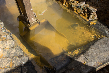 Rusty excavator shovel under dirty water from the sewerage system in the street. There is a bright light and shadows composing the scene