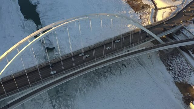 Aerial drop fly over Birds Eye View dolly roll the sunny lit white walter dale modern tie arch bridge with light traffic cracked iced North Saskatchewan River snow covered futuristic walkway design