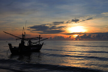 The silhouette of a fishing boat floats on the water in the morning, the sun is rising, the sky is beautiful blue and orange.