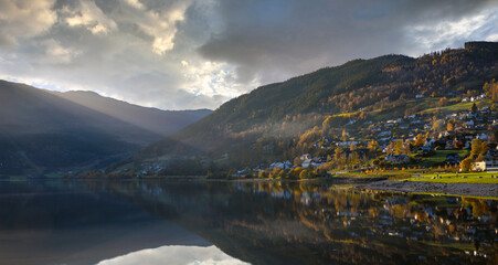 A panoramic view of the lake in Voss Norway, the water is still mirrored and the sun shines down in the evening, the house on the hill with beautiful trees in autumn.