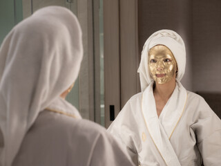 Obraz premium pretty woman in bathrobe with golden cosmetic mask does face massage near mirror in bathroom