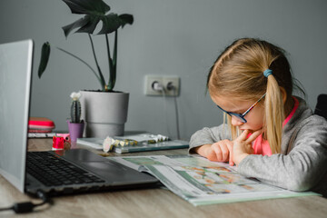 Little girl in eyelgasses looking at her classbook while having online lesson at home
