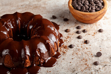 Angled view of a chocolate bundt cake with chocolate pudding icing dripping on top of it. There are cacao powder and chocolate chips on the surface with more chocolate chips in a wooden bowl in back.