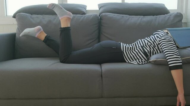 Young Asian girl lying on sofa feeling tired bored lazy sleeping book covering face fun kicking leg, tired late night study prepare for exam, living room quarantine teenage procrastination lifestyle.
