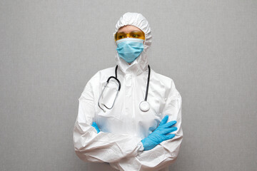 Fototapeta premium Woman doctor in protective suit, stethoscope, medical mask and yellow glasses with arms crossed on white background.