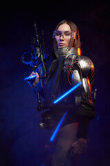 Naklejka premium Tattoed martial woman in cyberpunk style dressed in stylish black clothing. Fashionable and sexy female soldier holding a futuristic rifle and posing in dark background with blue lights.
