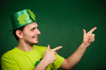 A man wearing a shamrock hat and a green T-shirt points his index fingers up. The guy is celebrating st patrick's day.