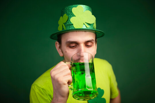 A man in a shamrock hat and green T-shirt drinks a large mug of green ale. Guy celebrates st patrick's day with a glass of beer. Studio photo