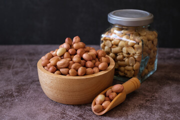 Raw Groundnuts in the wooden bowl and the wooden scoop with peanut snack in glass jar on cement background. 