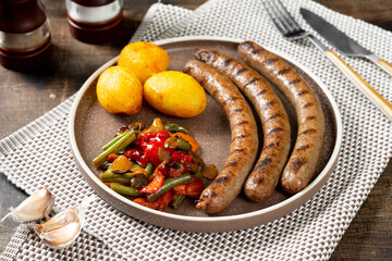 Grilled sausages. Grilled sausages with potatoes and pickled vegetables in a ceramic plate on a brown wooden table. Delicious meat sausages. Close-up