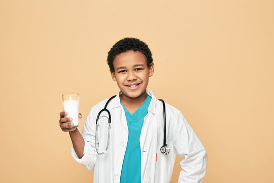 African American Boy Dressed Like A Doctor Holds A Glass Of Milk, On Beige Background. Benefits Of Milk For Kids