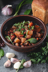 Bean soup with herbs in a clay bowl with fresh rye bread and garlic on a slate table on a dark background.
