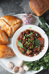 Bean soup with herbs in a white plate with fresh rye bread and garlic on a slate table on a dark background