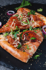 Slices Traditional Italian pepperoni pizza with tomatoes, cheese and flying onions and herbs, on a table of dark slate on a black background.