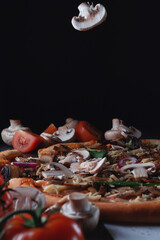 Pieces of flying fall mushrooms on Homemade Italian pizza on a cutting wooden board with ham, tomatoes, cheese, onions and herbs lies on a dark stone slate table