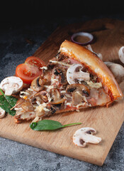 Slices of homemade Italian pizza on a chopping wooden board with mushrooms, ham, tomatoes, cheese, onions and herbs lie on a dark stone slate table