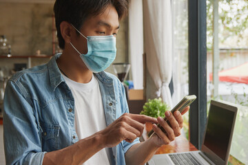 Fototapeta na wymiar Zoom View Front Right Asian Casual Businessman in Denim or Jeans Shirt Wear Face Mask Touch Smartphone in Coffee Shop. Casual Businessman with Technology in Covid 19 Situation