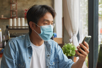 Zoom View Front Right Asian Casual Businessman in Denim or Jeans Shirt Wear Face Mask Using Smartphone in Coffee Shop. Casual Businessman with Technology in Covid 19 Situation