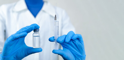 Female doctor in a white coat, mask and blue gloves holds a syringe in one hand, and in the other a bottle of medicine close-up, copy the space. Banner. Concept of medicine, medicines, and vaccination