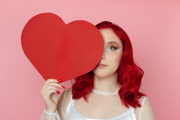 close-up serious, neutral young woman with red hair hides half of her face behind a large paper red heart isolated on a pink background.