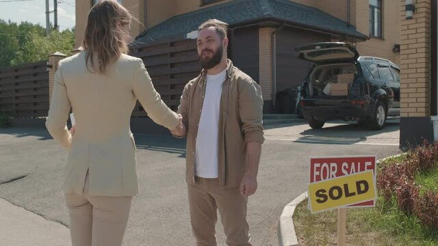  Dolly-in shot of young female real estate agent hanging house keys to happy bearded man and shaking his hand