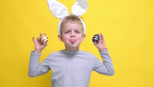 Portrait Easter kid. Boy in rabbit bunny ears on head having fun looking into the camera sticks out tongue making crazy funny face with colored Easter eggs on his eyes yellowfins studio background.