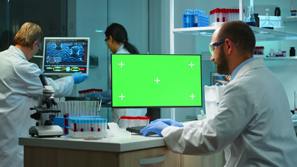 Man researcher looking at chroma key computer in modern equipped lab working overtime. Team of microbiologists doing vaccine research writing on device with green screen, isolated, mockup display.