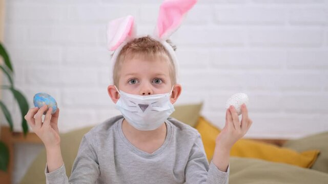 Boy in rabbit bunny ears on head and protective mask having fun looking into the camera making crazy funny face with colored Easter eggs on his eyes at home. Cheerful smiling child. Covid easter kid