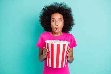 Photo portrait of amazed black skin girl holding bucket of popcorn isolated on vivid cyan colored background - obrazy, fototapety, plakaty
