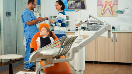 Nurse inviting kid patient in consultation dental room, pediatric doctor speaking with little girl. Man assistant talking with mother preparing for stomatologic examination in orthodontist office